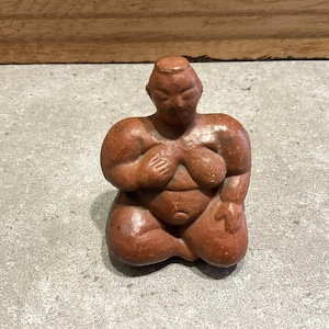 Puede incluir: Pequeña figura de color marrón rojizo de una figura sentada con rasgos exagerados. La figura tiene senos grandes y un vientre redondeado, con los brazos apoyados en las rodillas. La escultura probablemente está hecha de arcilla o un material similar.