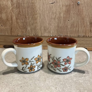 May include: Two ceramic mugs with white bodies, brown rims, and handles. Each mug features a floral design in orange and brown. The mugs are set against a wooden background.