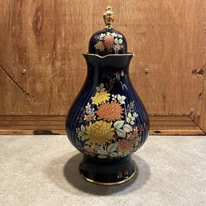 May include: A dark blue ceramic ginger jar with a matching lid featuring a gold-coloured finial. The jar has a ribbed body decorated with a floral pattern of yellow, orange, and white chrysanthemums and green leaves, finished with a gold rim.