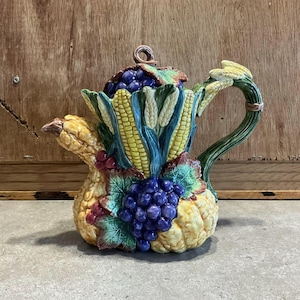 Peut inclure: Une théière en céramique en forme de corne d'abondance avec du maïs jaune, des feuilles vertes, des raisins violets et une gourde jaune. La théière a une poignée brune et un couvercle avec un bouton brun.