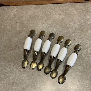 以下が含まれることがあります： 真鍮製のベースに白いセラミック製のキャビネットノブが6個あります。ノブは楕円形で、シンプルなデザインです。