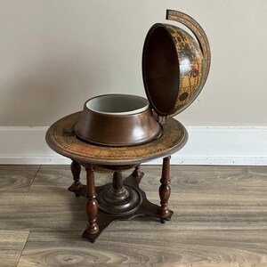 May include: A vintage globe bar with a hinged top, revealing a white interior. The globe sits on a round table with a map design and four legs. The bar is made of wood and has a dark brown finish.