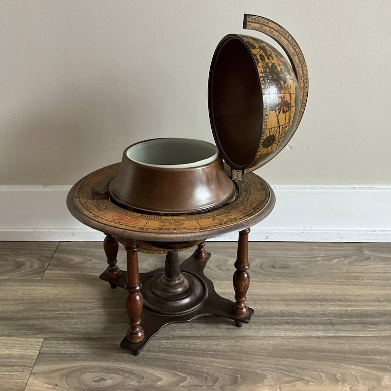 May include: A vintage globe bar with a hinged top, revealing a white interior. The globe sits on a round table with a map design and four legs. The bar is made of wood and has a dark brown finish.