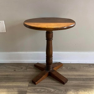 May include: A round wooden side table with a brown finish. The table has a single pedestal base with a cross-shaped foot.