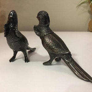 May include: Two silver metal bird figurines with detailed feather patterns. The birds are facing each other with their heads turned slightly to the side.