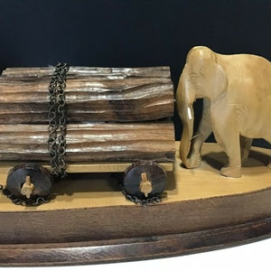 May include: A wooden elephant figurine pulling a cart loaded with logs. The cart has two wheels and is attached to the elephant by a chain. The figurine is on a wooden base.