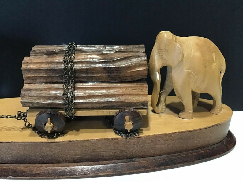 May include: A wooden elephant figurine pulling a cart loaded with logs. The cart has two wheels and is attached to the elephant by a chain. The figurine is on a wooden base.
