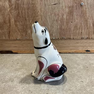 Könnte beinhalten: Eine weiße Keramik-Hundefigur mit schwarzen und rosafarbenen Details. Der Hund sitzt mit dem Kopf nach oben geneigt und leicht geöffnetem Maul. Die Figur hat ein schwarzes Halsband und einen rosafarbenen Fleck auf dem Rücken.