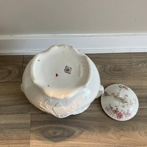 May include: White porcelain serving bowl with lid. The bowl has a scalloped edge and ornate relief detailing. The lid features pink floral and gold accents. The underside of the bowl has a red mark and a maker's mark.