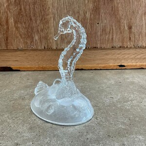 May include: A clear glass sculpture of a seahorse with a textured surface. The seahorse is standing on a base with a textured surface.