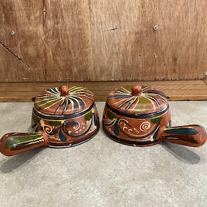 Peut inclure: Deux bols à couvercle en céramique peints à la main avec des poignées. Les bols sont principalement en terre cuite avec des motifs floraux et tourbillonnants colorés en vert, bleu et blanc. Les couvercles ont un petit bouton sur le dessus.