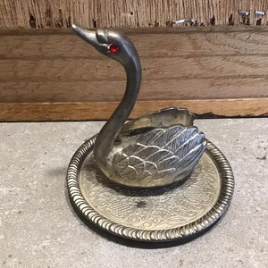 Puede incluir: Porta anillos de cisne plateado con un ojo de joya roja, sentado en una bandeja de plata decorativa con un diseño ornamentado.