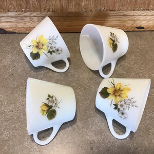 Pode incluir: Quatro canecas de cer&acirc;mica branca com desenhos florais amarelos e brancos. As canecas t&ecirc;m uma forma arredondada e uma al&ccedil;a lateral.