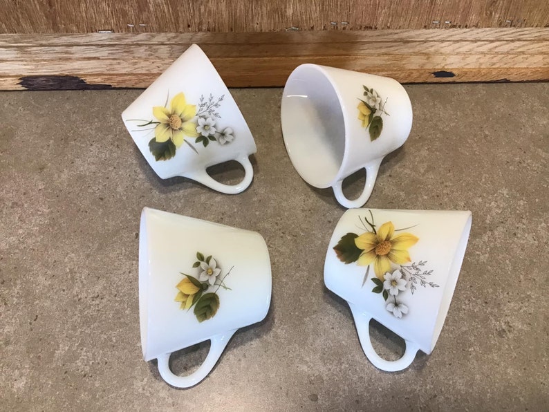 Pode incluir: Quatro canecas de cer&acirc;mica branca com desenhos florais amarelos e brancos. As canecas t&ecirc;m uma forma arredondada e uma al&ccedil;a lateral.