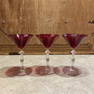 May include: Three vintage martini glasses with ruby red bowls and clear, ornate stems. The glasses have a classic, triangular shape and sit on round bases. The stems feature a twisted design. The background is a wooden surface.