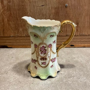 Pode incluir: Um jarro de porcelana vintage com alça dourada e design floral. O jarro tem um fundo verde claro e branco com flores rosa e vermelhas.