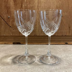 May include: Two clear crystal wine glasses with etched designs. The glasses have a slender stem and a round base. The etched design features a series of diagonal lines and curves. The glasses are displayed on a gray surface against a wooden background.