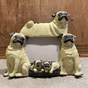 Puede incluir: Un marco de fotos beige con forma de perro pug con cuatro figuras de perros pug. Dos pugs se sientan a cada lado del marco, un pug se acuesta en la parte superior y tres cachorros pug están en una canasta en la parte inferior.