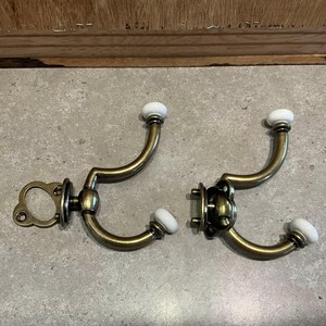 Puede incluir: Dos perchas de pared antiguas de color latón con pomos de cerámica blanca. Los ganchos tienen un diseño curvo y están montados sobre placas traseras decorativas. Los ganchos están diseñados para colgar objetos y añaden un toque vintage.