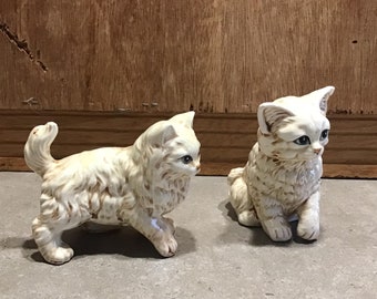 May include: Two ceramic white kittens with brown markings. One kitten is sitting with its head turned to the side, while the other is standing with its head turned to the left. Both kittens have blue eyes.