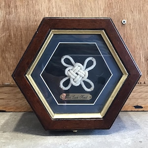 Op de afbeelding: Een ingelijst, wit touwknot in een zeshoekige houten doos met een gouden rand. De knoop is gelabeld "Good Luck Knot".