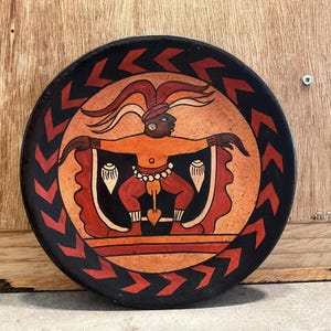 May include: A round ceramic plate with a black background and a red and orange design. The design features a stylized figure with a headdress and a red and orange skirt. The figure is surrounded by a black and red chevron pattern.