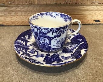 May include: A blue and white porcelain teacup and saucer set with a gold trim. The set features a traditional Japanese design with a pagoda and figures in blue.