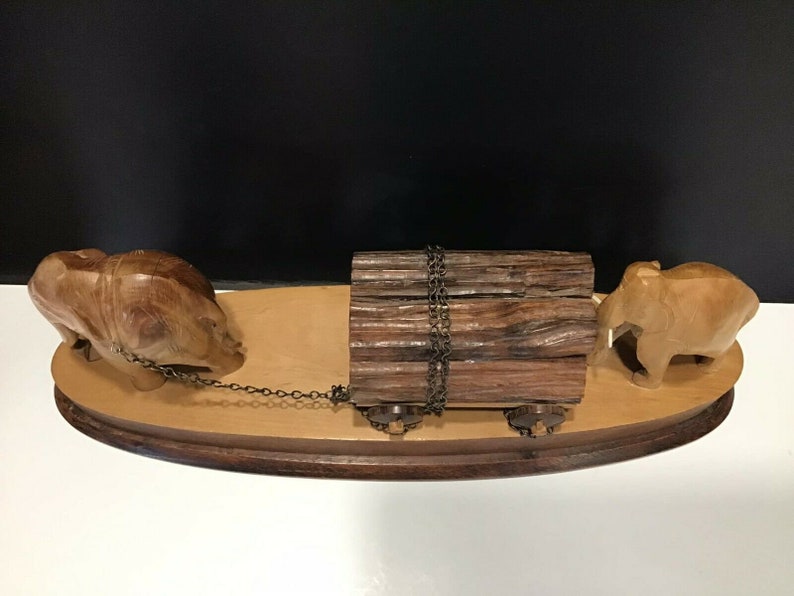 May include: A wooden sculpture of two elephants pulling a log on a wooden base. The elephants are carved in a realistic style and the log is tied with a chain.
