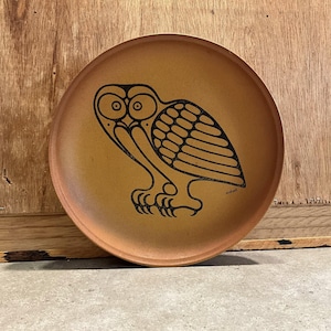 May include: A round, brown plate with a black owl illustration. The owl is stylized with large eyes and a detailed body. The plate has a slightly raised edge and a matte finish, suitable for decorative use.