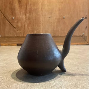 May include: A dark brown, matte-finished ceramic vessel with a wide, rounded base that tapers toward a circular opening at the top. The object features a single, elongated, curved handle extending outward from the side, creating a distinct silhouette.