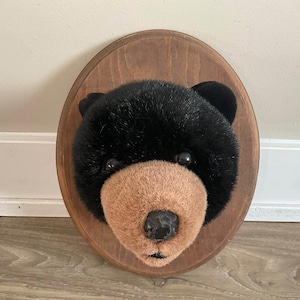 May include: A plush black bear head wall mount featuring soft black fur, a tan muzzle, and a black nose. The bear head is securely attached to a smooth, oval-shaped wooden plaque with a medium brown finish, designed for display on a vertical surface.