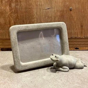 May include: A grey stone picture frame with a frog figurine sitting in front of it. The frame has a textured surface and a rectangular shape.