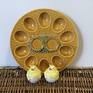 Pode incluir: Um prato de ovos de cerâmica amarelo com 10 reentrâncias individuais em forma de ovo. O prato tem um design verde e marrom no centro. Dois saleiros e pimenteiros de cerâmica amarela em forma de pintinho estão em primeiro plano.