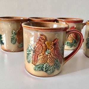 May include: Set of six ceramic mugs with a brown and beige glaze. Each mug features a raised design of cardinals and pine branches.
