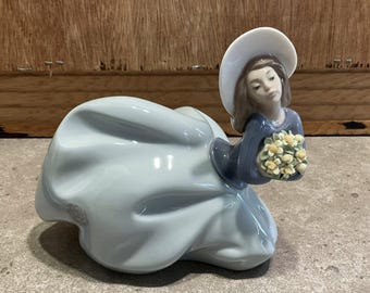 May include: A porcelain figurine of a woman in a light blue dress and a white hat, holding a bouquet of yellow flowers. The figurine has a serene expression and is set against a neutral background.