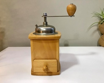 Puede incluir: Un molinillo de café de madera con tapa y mango de acero inoxidable. El molinillo tiene un pequeño cajón y un pomo de madera en el mango. La madera es de color claro.