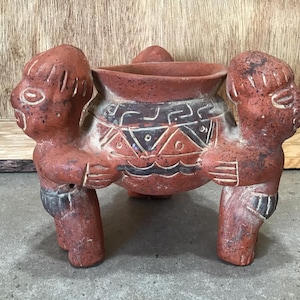 Peut inclure: Un pot en terre cuite porté par trois personnages. Le pot est décoré de motifs géométriques en noir et blanc. Les personnages ont des visages stylisés et portent des vêtements simples.