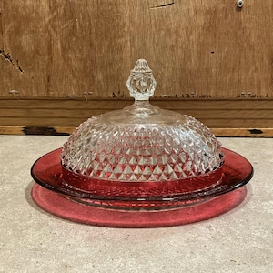 May include: A vintage red glass butter dish with a clear, diamond-patterned lid. The lid has a decorative finial. The dish sits on a matching red base, creating a classic and elegant look. This item is perfect for serving butter.