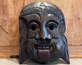 May include: A dark brown wooden mask with a carved face, a wide mouth, and a protruding tongue. The mask has a triangular design on the forehead and intricate details around the eyes.