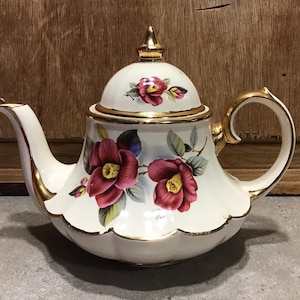 Puede incluir: Tetera blanca con ribete dorado y diseño floral con camelias rosas. La tetera tiene una tapa abovedada con un pomo dorado.