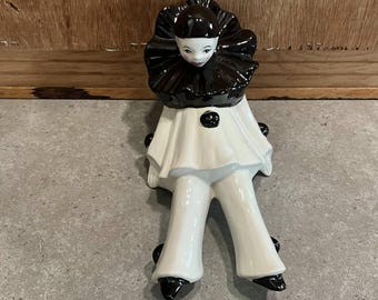 Puede incluir: Figura de cerámica de un payaso Pierrot sentado. La figura tiene un cuello negro con volantes y un atuendo blanco con detalles negros. La cara del payaso es pálida con rasgos delicados. Un artículo decorativo.