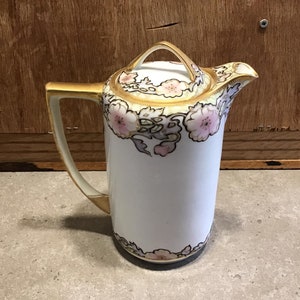 Peut inclure: Théière en porcelaine blanche avec un motif floral rose, or et vert. La théière a un couvercle et une poignée.