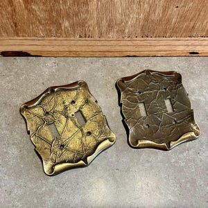 Op de afbeelding: Twee gouden lichtschakelaars met een getextureerd, reliëfdesign. De schakelaars hebben een decoratief, versierd frame.
