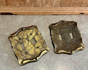 May include: Two gold-toned light switch covers with a textured, embossed design. The covers have a decorative, ornate frame.