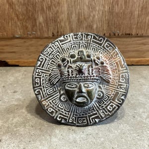 May include: A circular, gray and brown ceramic sculpture of a stylized face with a feathered headdress. The face has a wide mouth and large eyes. The sculpture is surrounded by a geometric pattern.