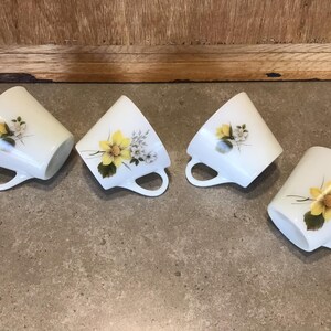 Pode incluir: Quatro ch&aacute;venas de ch&aacute; de cer&acirc;mica branca com desenhos florais amarelos e brancos. As ch&aacute;venas est&atilde;o dispostas em fila sobre uma superf&iacute;cie cinzenta.