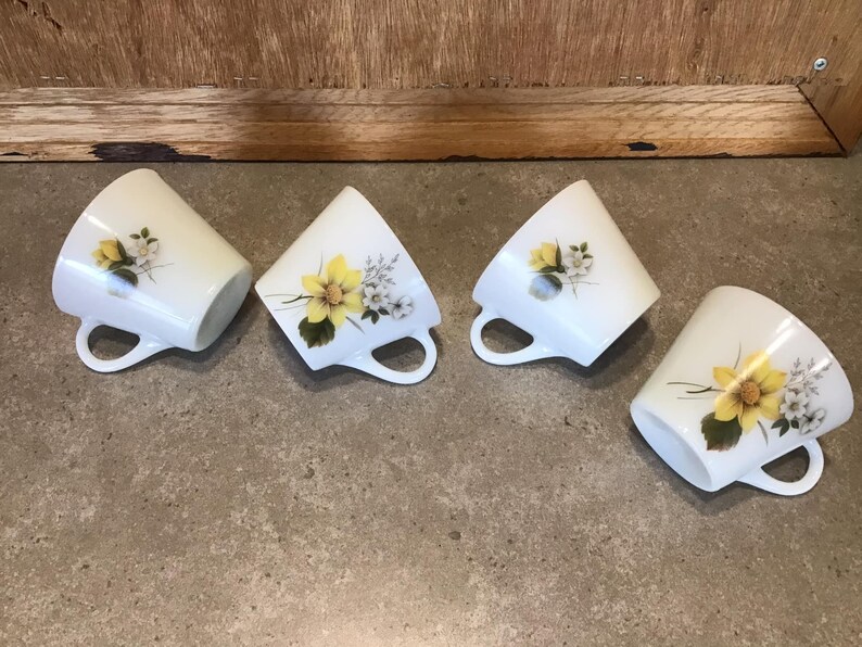 Pode incluir: Quatro ch&aacute;venas de ch&aacute; de cer&acirc;mica branca com desenhos florais amarelos e brancos. As ch&aacute;venas est&atilde;o dispostas em fila sobre uma superf&iacute;cie cinzenta.
