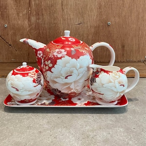 Peut inclure: Un service à thé composé d'une théière, d'un sucrier avec couvercle et d'un pot à lait sur un plateau rectangulaire. Chaque pièce est ornée d'un fond rouge vif, de grands motifs floraux blancs et de détails dorés complexes.