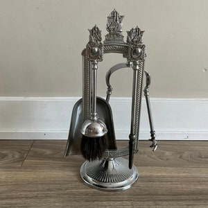 Puede incluir: Conjunto de herramientas para chimenea de plata con cepillo, pala y atizador. Las herramientas están montadas en un soporte con un diseño de barco y las palabras "Mayflower" en la parte superior.