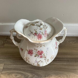 May include: A white porcelain chamber pot with a lid, adorned with pink floral patterns and gold accents. The pot features two handles and a scalloped rim, showcasing a vintage aesthetic.